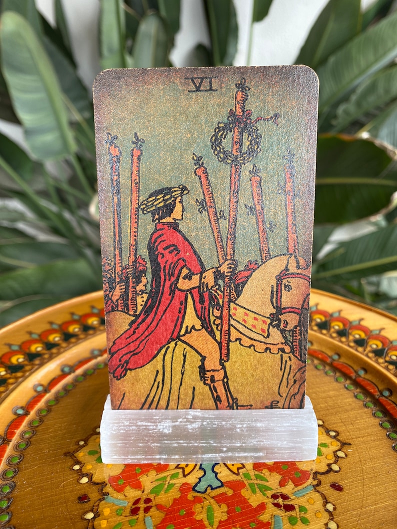 May include: A vintage tarot card with a worn, aged look. The card depicts a man on horseback, holding a staff. The card number VII is visible in the top right corner. The card is on a white stand.