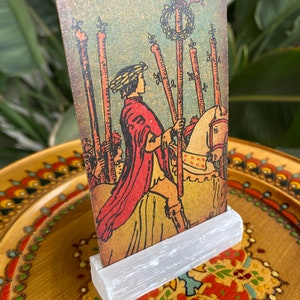 May include: A vintage tarot card depicting the VII of Wands card. The card features a man on horseback holding a staff, with five other staffs standing upright in the background. The card is set against a blue and green background with a wreath of flowers on top of the staffs.