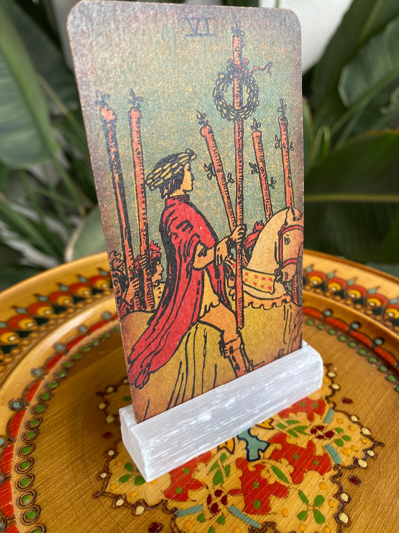 May include: A vintage tarot card depicting the VII of Wands card. The card features a man on horseback holding a staff, with five other staffs standing upright in the background. The card is set against a blue and green background with a wreath of flowers on top of the staffs.