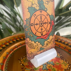May include: A tarot card with a wheel of fortune design. The card is yellow, orange, red, green, and blue. The card is titled "Wheel & Fortune". The card is standing on a white acrylic card holder.