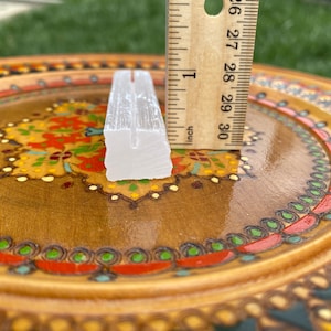 May include: A white selenite crystal wand measuring approximately 2 inches in length. The wand is sitting on a wooden plate with colorful painted designs.