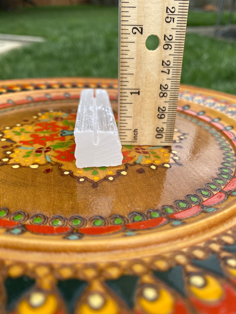 May include: A white selenite crystal wand measuring approximately 2 inches in length. The wand is sitting on a wooden plate with colorful painted designs.