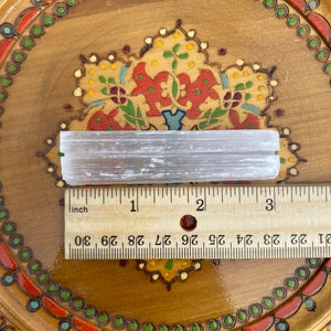 May include: A white selenite stick measuring approximately 3 inches long, resting on a wooden surface with a decorative pattern. The stick is smooth and has a natural, crystalline texture.