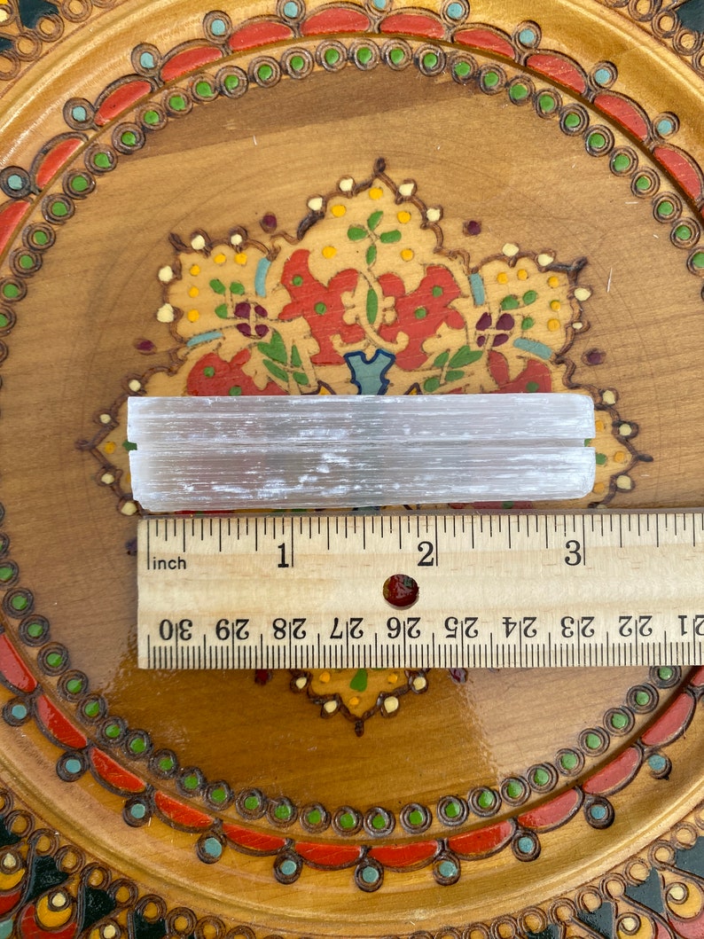 May include: A white selenite stick measuring approximately 3 inches long, resting on a wooden surface with a decorative pattern. The stick is smooth and has a natural, crystalline texture.