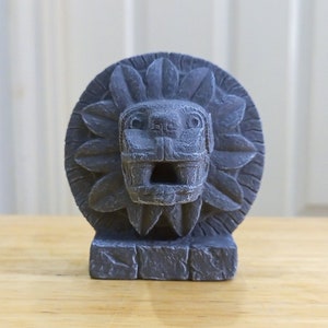 Könnte beinhalten: Eine graue Steinskulptur eines stilisierten Tierkopfes mit einem kreisförmigen Design darum herum. Die Skulptur steht auf einem kleinen grauen Steinsockel.