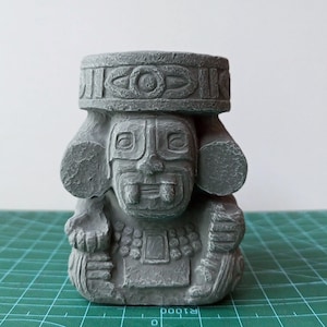 May include: A gray, Aztec-style statue with a detailed headdress and facial features. The sculpture has a rectangular body and is sitting on a green cutting mat. The statue is a decorative item.