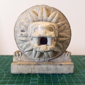 Op de afbeelding: Een wit stenen beeld van een mythisch wezen met een grote, open mond en een zon-achtig ontwerp erachter. Het beeld staat op een rechthoekige basis.