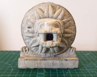 May include: A white stone sculpture of a mythical creature with a large, open mouth and a sun-like design behind it. The sculpture is on a rectangular base.
