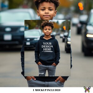 Puede incluir: Un joven lleva una sudadera con capucha azul marino con el texto blanco "YOUR DESIGN HERE". La sudadera tiene un bolsillo delantero y una capucha con cord&oacute;n. La persona lleva vaqueros grises. El fondo muestra una calle con coches.