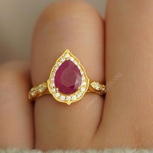 Op de afbeelding: Een gouden ring met een peervormige, dieprode edelsteen, omringd door kleine, heldere stenen. De ringband heeft een delicaat, sierlijk ontwerp. De ring is geplaatst tegen een zachte, neutrale achtergrond.