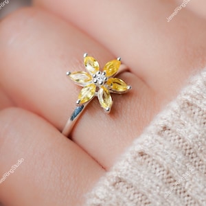 以下が含まれることがあります： 黄色い花のデザインが施された銀の指輪。花は6枚の花びらがあり、中央に小さな透明な石が埋め込まれています。