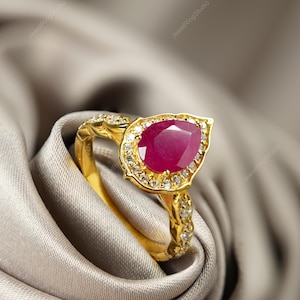 Op de afbeelding: Een gouden ring met een peervormige, dieprode edelsteen in het midden, omringd door kleinere, heldere stenen. De band heeft ingewikkelde details en is geplaatst tegen een zachte, neutraal getinte stof.