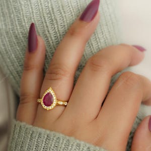 Op de afbeelding: Een gouden ring met een peervormige, dieprode edelsteen, omringd door kleine, heldere stenen. De ring zit op een gouden band met meer kleine stenen. De ring zit aan een vinger, met een lichtgroene trui op de achtergrond.