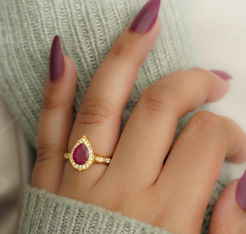 Op de afbeelding: Een gouden ring met een peervormige, dieprode edelsteen, omringd door kleine, heldere stenen. De ring zit op een gouden band met meer kleine stenen. De ring zit aan een vinger, met een lichtgroene trui op de achtergrond.