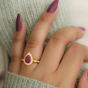 Op de afbeelding: Een gouden ring met een peervormige, dieprode edelsteen, omringd door kleinere, heldere stenen. De ring is gezet op een gouden band met extra kleine stenen. De ring wordt getoond op een hand met gelakte nagels.