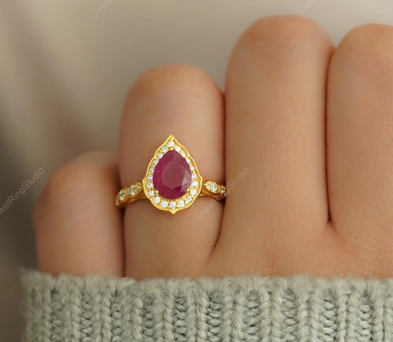 Op de afbeelding: Een gouden ring met een peervormige, dieprode edelsteen, omringd door kleine, heldere stenen. De ringband heeft een delicaat, sierlijk ontwerp. De ring is geplaatst tegen een zachte, neutrale achtergrond.