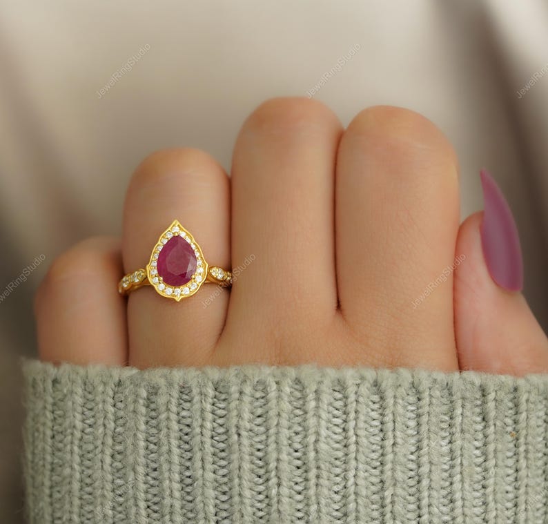 Op de afbeelding: Een gouden ring met een peervormige, dieprode robijn als middensteen, omringd door kleine witte edelstenen. De band van de ring heeft een delicaat, sierlijk ontwerp. De ring is geplaatst tegen een zachte, neutrale achtergrond.