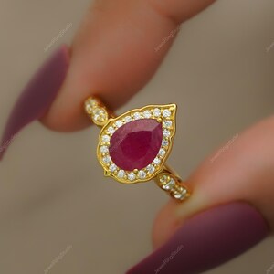Op de afbeelding: Een gouden ring met een peervormige, dieprode edelsteen, omringd door kleine, heldere stenen. De ring heeft een gouden band en zetting, met extra kleine stenen op de band. De ring wordt tussen twee vingers gehouden.