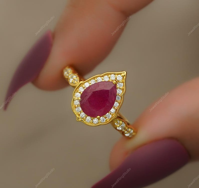 Op de afbeelding: Een gouden ring met een peervormige, dieprode edelsteen, omringd door kleine, heldere stenen. De ring heeft een gouden band en zetting, met extra kleine stenen op de band. De ring wordt tussen twee vingers gehouden.