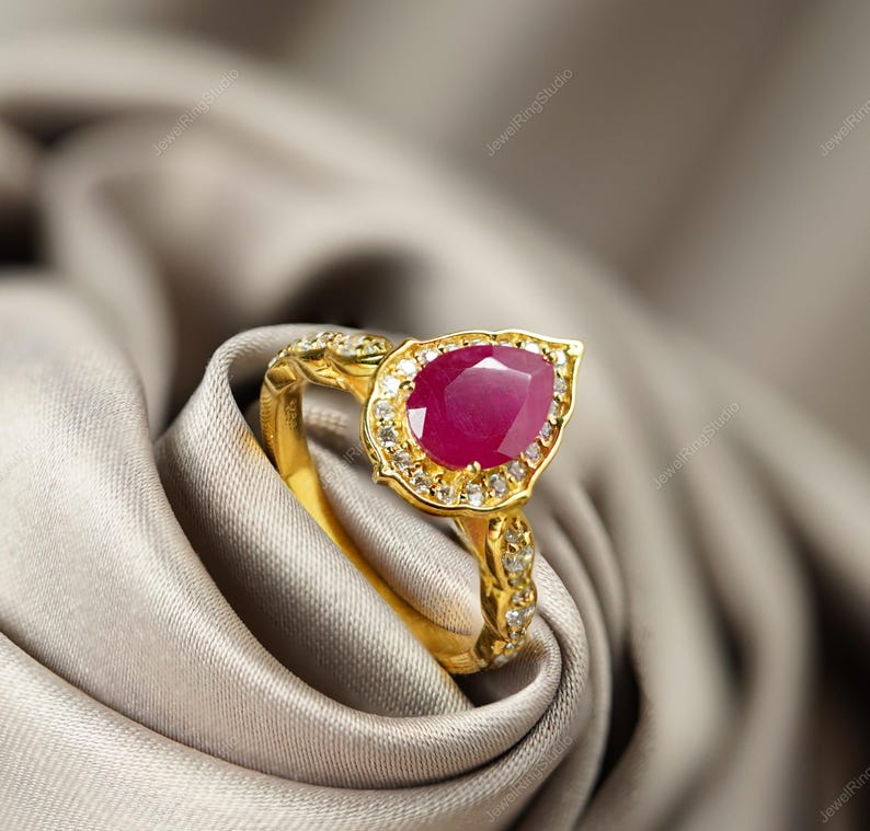 Op de afbeelding: Een gouden ring met een peervormige, dieprode edelsteen in het midden, omringd door kleinere, heldere stenen. De band heeft ingewikkelde details en is geplaatst tegen een zachte, neutraal getinte stof.