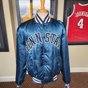 Puede incluir: Una chaqueta de béisbol de satén azul con ribetes de canalé blanco y azul marino. La chaqueta tiene un logotipo bordado en azul marino "Penn State" en la parte delantera.