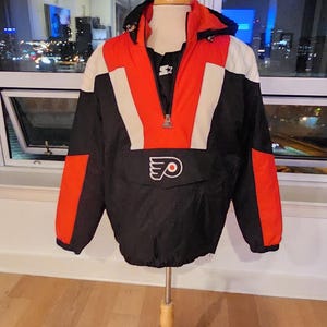 Peut inclure: Veste Starter des Flyers de Philadelphie noire, rouge et blanche, présentée sur un mannequin en bois. La veste arbore le logo des Flyers et un logo Starter. En arrière-plan, une vue nocturne de la ville.