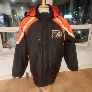 Könnte beinhalten: Schwarze und orangefarbene Winterjacke mit dem Logo der Philadelphia Flyers auf der Brust. Die Jacke hat eine Kapuze, einen Reißverschluss und orangefarbene und weiße Streifen an den Ärmeln. Die Jacke wird auf einer Schaufensterpuppe präsentiert.