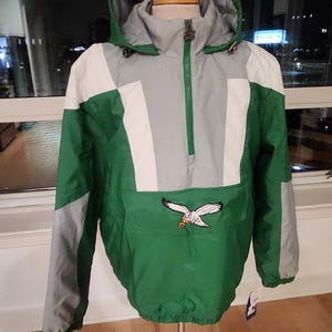 Pode incluir: Casaco anoraque verde, cinzento e branco com capuz e bolso frontal. O casaco tem fecho de correr e um logótipo de águia. É exibido num manequim de madeira. Medidas em cm.
