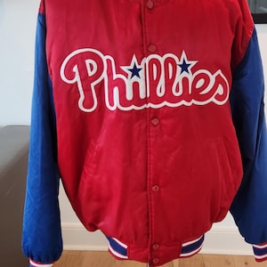 Peut inclure: Une veste de baseball en satin rouge avec des manches bleues et des bordures blanches. La veste a un logo "Phillies" blanc avec deux étoiles sur le devant.