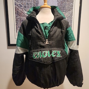 Op de afbeelding: Een zwarte, groene en zilverkleurige pulloverjas met een halve ritssluiting en een Starter-logo als ritstrekker. Het woord EAGLES is in groen en wit geborduurd op de klep van de voorzak. De jas heeft een hoge kraag en lange mouwen.