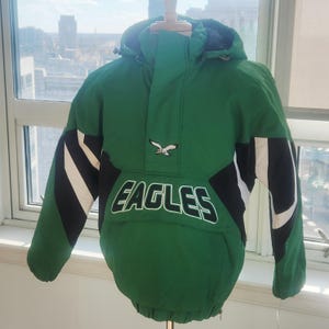 May include: Green and black hooded anorak jacket with the word "EAGLES" in white block letters. The jacket has a white and black design on the sleeves and an eagle emblem. The jacket is displayed on a mannequin.