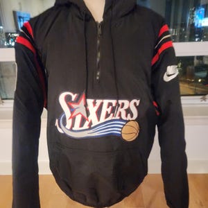 Könnte beinhalten: Schwarze Anorakjacke mit Kapuze und dem Logo der Philadelphia 76ers auf der Vorderseite. Die Jacke hat rote und weiße Streifen auf den Ärmeln und ein weißes Nike-Logo. Sie hat einen halben Reißverschluss und eine Vordertasche.