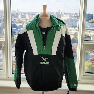 May include: A black, green, and white hooded anorak jacket. The jacket features a green and white chevron design on the front, with the team logo and the word "Eagles" on the front pocket. The jacket has a zipper closure and a Starter logo.