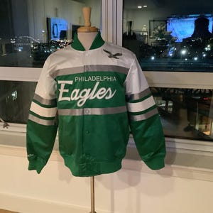 Op de afbeelding: Een groene en witte satijnen jas met de tekst "PHILADELPHIA Eagles" in witte letters. De jas heeft een groene body, witte mouwen en zilveren strepen. Een kleine adelaarsafbeelding staat boven de teamnaam. De jas wordt op een mannequin getoond.