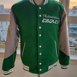 Può includere: Giacca da baseball verde e grigia con lettering bianco che dice "Philadelphia Eagles". La giacca è su un manichino di legno.