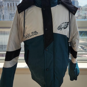 Könnte beinhalten: Eine Vintage-Jacke der Philadelphia Eagles, ausgestellt auf einer Holzpuppe. Die Jacke hat ein Farbblockdesign in Teal, Weiß und Schwarz mit dem Eagles-Logo und dem Teamnamen. Sie hat eine Kapuze und eine Knopfleiste.