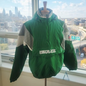 May include: Green and gray pullover jacket with the word "EAGLES" in white across the front. The jacket has a hood and a small NFL logo on the sleeve. The jacket is displayed on a mannequin.