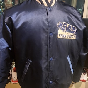 Puede incluir: Una chaqueta bomber de satén azul marino con ribetes de canalé blanco y azul marino en el cuello, los puños y el dobladillo. La chaqueta tiene cierre de botones y presenta un logotipo bordado de Penn State con un león en el pecho izquierdo.