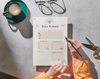 Op de afbeelding: Een Daily Planner-blad met een zon- en oogontwerp, met secties voor takenlijsten, zelfzorg en affirmaties. Een persoon schrijft met een potlood op de planner. Een turquoise kopje koffie en een gouden schaar liggen ook op tafel.
