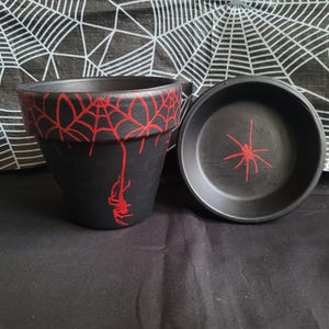 Puede incluir: Juego de maceta y platillo de cerámica negra con diseños de telarañas y arañas rojas. La maceta tiene un diseño de telaraña roja alrededor del borde y una araña colgando a un lado. El platillo tiene un diseño de araña roja en el centro.