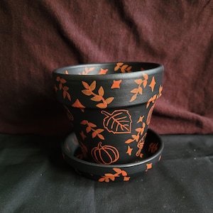 May include: Black ceramic flower pot with matching saucer, decorated with orange leaf and pumpkin designs. The pot has a classic shape, perfect for indoor or outdoor plants. A seasonal, decorative item.