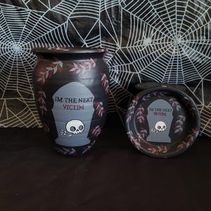 May include: A black ceramic vase and small dish with a Halloween theme. Both items feature a tombstone design with the text "IM THE NEXT VICTIM" and a skull. The vase and dish are decorated with red and white leaf accents.