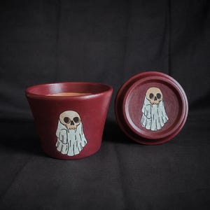 May include: A dark red ceramic pot and a matching circular lid, both featuring a hand-painted illustration of a skull wearing a white ghost sheet. The pot has a tapered shape, and the lid is positioned to the right, displaying the same ghost motif.