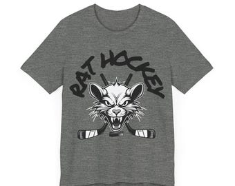 Op de afbeelding: Een grijs t-shirt met een afbeelding van een woeste rat met hockeysticks en de tekst "RAT HOCKEY" in het zwart. De rat heeft een boze uitdrukking. Het shirt heeft een ronde hals en korte mouwen.