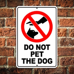May include: White sign with a red circle and a diagonal line through it. Inside the circle is a black silhouette of a dog's head. The text "DO NOT PET THE DOG" is printed in black letters below the circle.