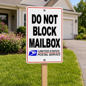 May include: A rectangular white sign with a red border and the words "DO NOT BLOCK MAILBOX" in bold black letters. The United States Postal Service logo is at the bottom. The sign is mounted on a wooden post in a grassy yard.