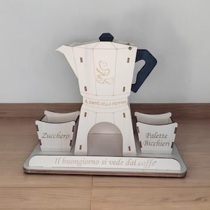 Può includere: Stendino in legno bianco per caffettiera con due scomparti etichettati "Zucchero" e "Palette Bicchieri". Lo stendino presenta un'apertura per una caffettiera e la scritta "Il buongiorno si vede dal caffe".