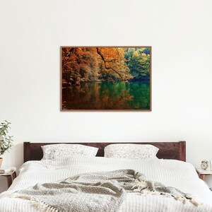 May include: A framed photograph of a lake surrounded by trees with fall foliage. The water is still and reflects the trees and sky. The colors are vibrant and warm, with shades of orange, yellow, green, and blue.