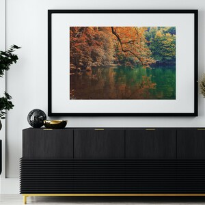 May include: A framed photograph of a lake surrounded by trees with fall foliage. The lake is reflecting the colors of the trees. The frame is black with a white mat.