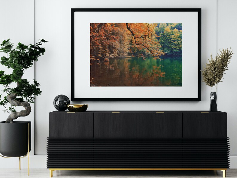 May include: A framed photograph of a lake surrounded by trees with fall foliage. The lake is reflecting the colors of the trees. The frame is black with a white mat.